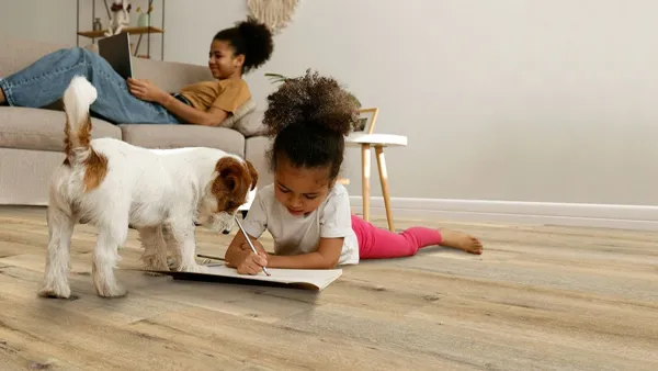 In this cozy living room scene, a girl writes on the floor (with a dog beside her) flooring with excellent abrasion resistance