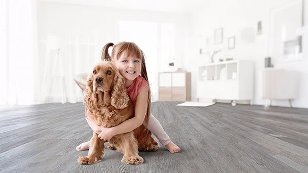Una niña se sienta con las piernas cruzadas sobre el suelo SPC resistente a los arañazos y a prueba de mascotas, acariciando a su juguetón perro.
