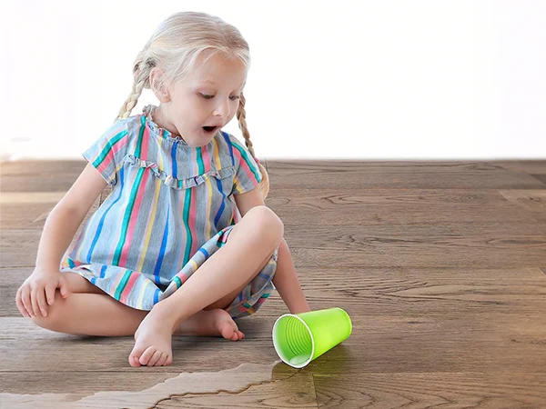 A little girl sits on SPC flooring, playfully splashing water from a green cup.